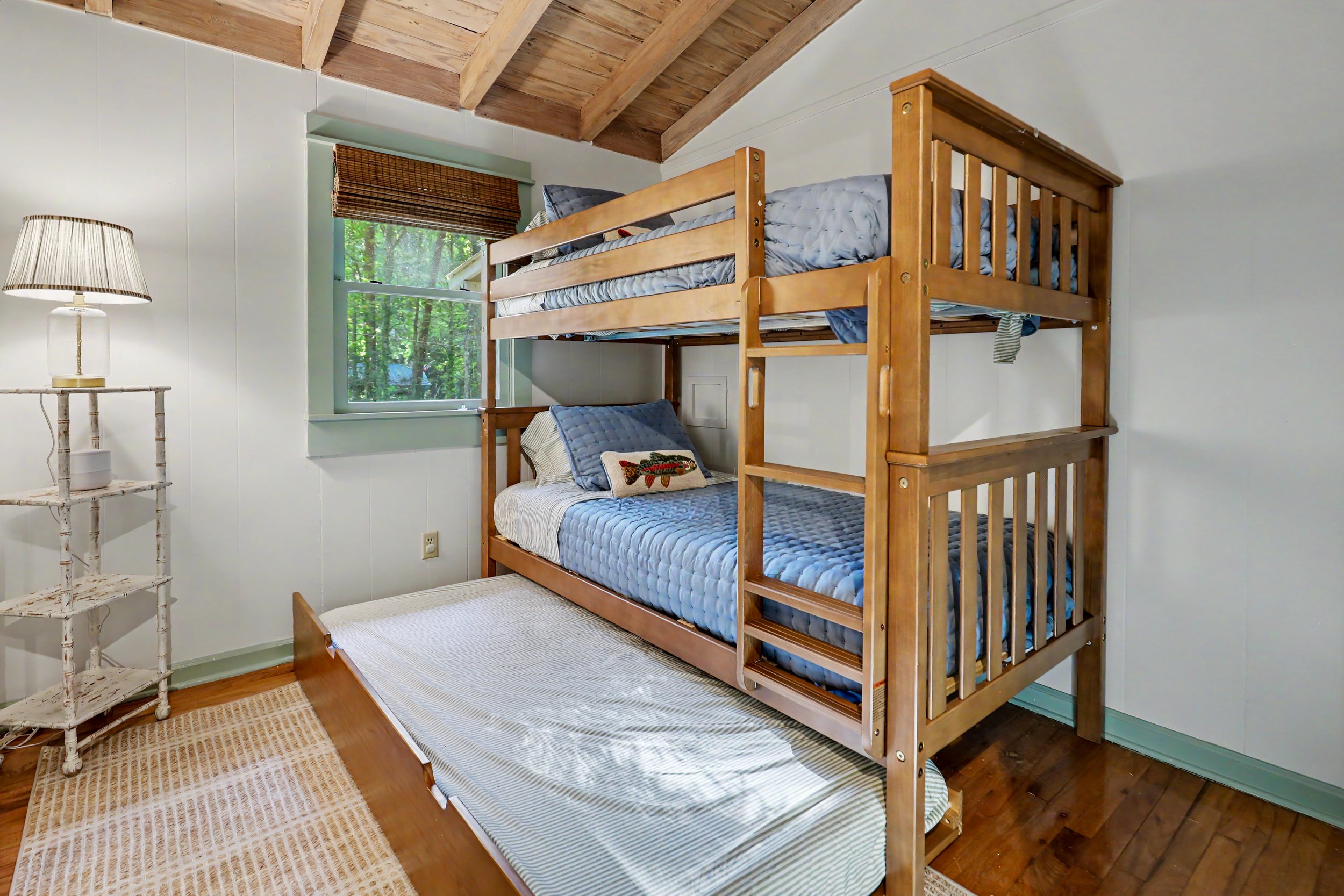 A family-friendly Lake Burton cabin rental bedroom with a wooden bunk bed, featuring mattresses and pillows, sunlight coming through a window, a white side table with a lamp, a woven rug, and wood-paneled ceiling.