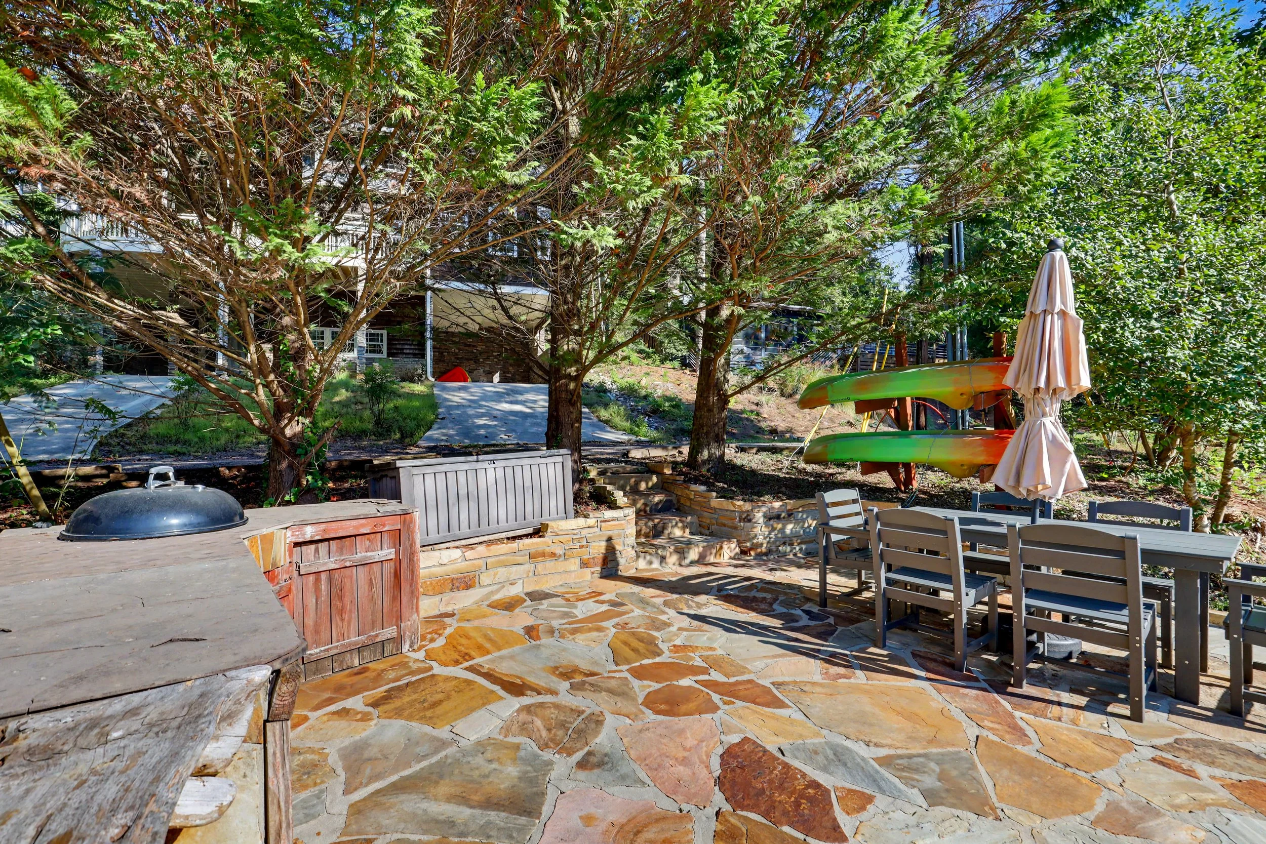 Backyard patio on Lake Burton with stone flooring, outdoor table with chairs, a large tree providing shade, two colorful kayaks stacked on a stand, a closed umbrella, and a grill.