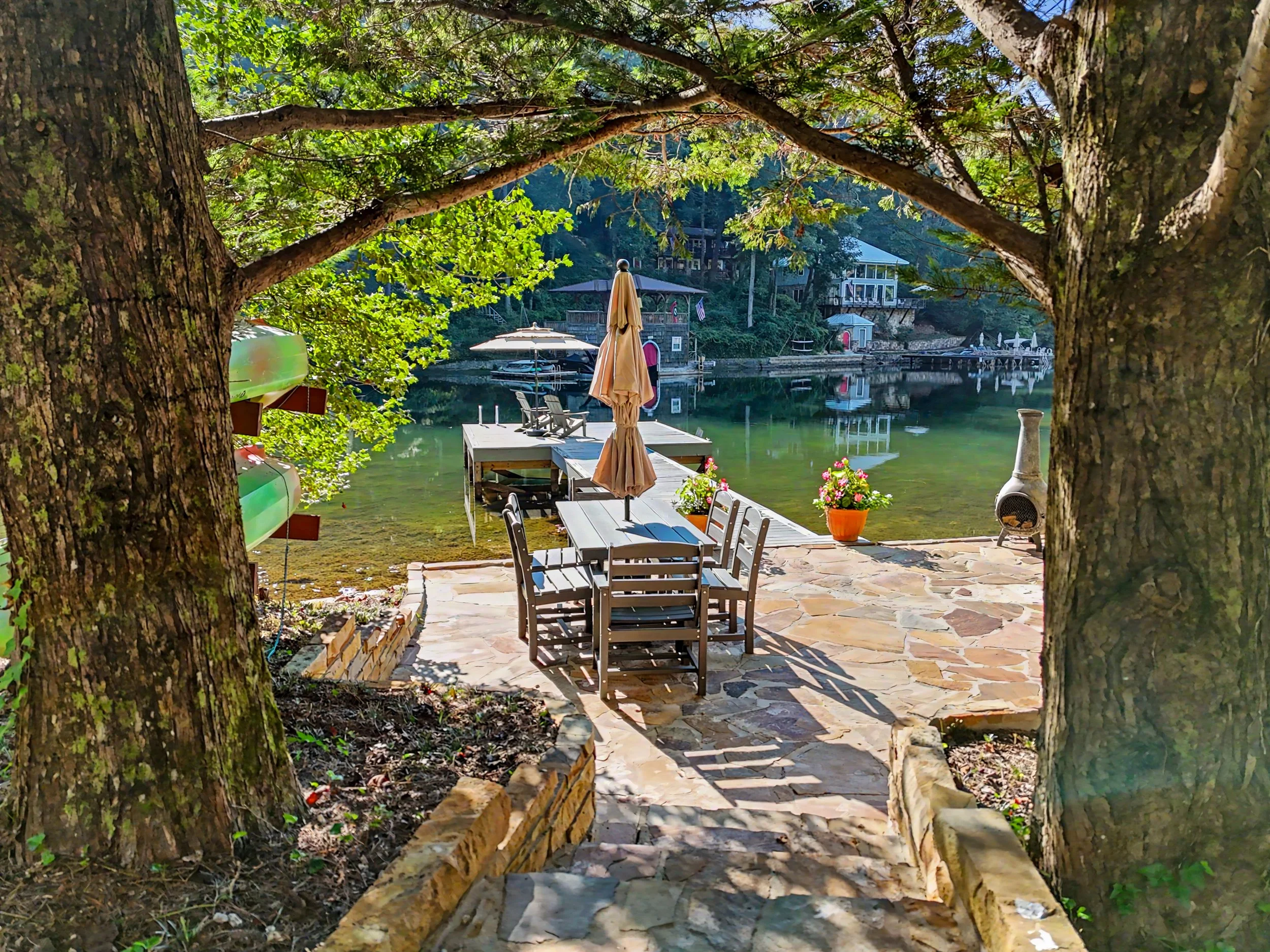 A lakeside outdoor patio on Perrin Cove at Lake Burton with tables, chairs, and closed umbrellas, surrounded by trees and houses, reflecting on the water.