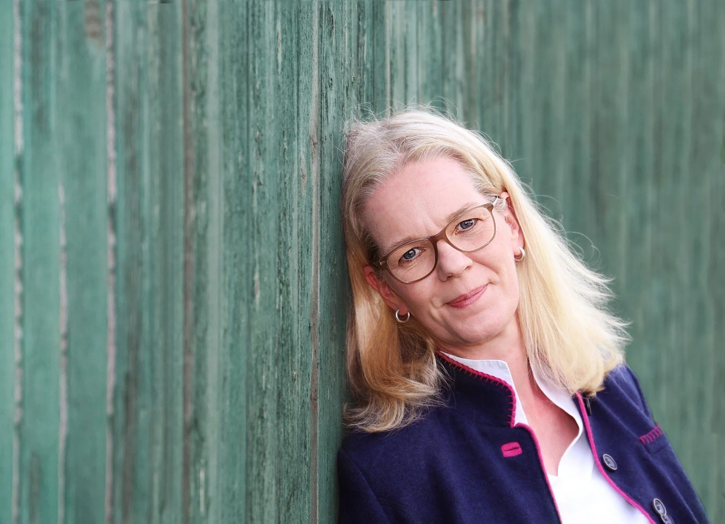 Eine blonde Frau mit Brille, die an einer grünen Holzwand lehnt.