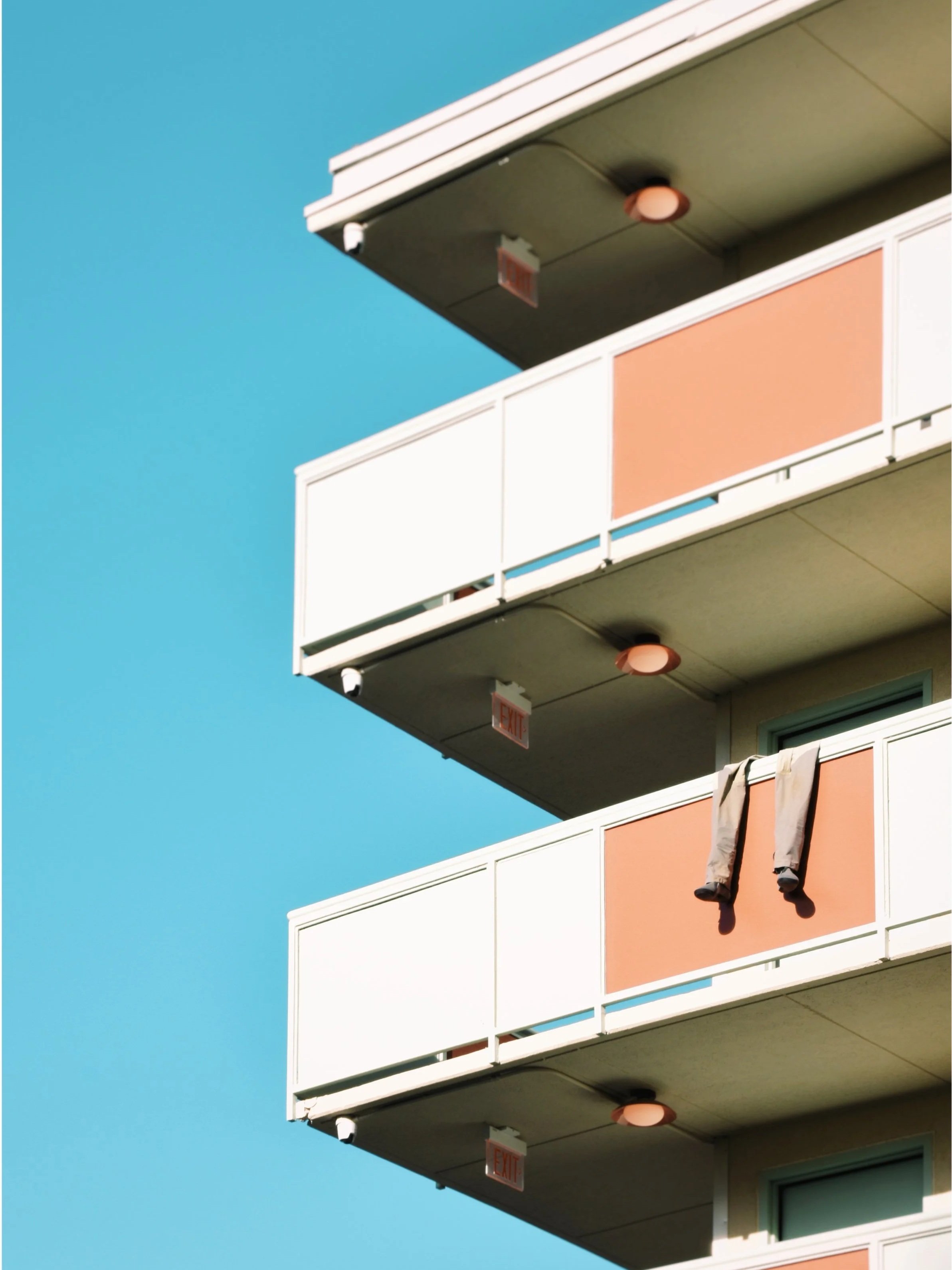 Three balconies of a modern apartment building against a clear blue sky, with clothing hanging on one balcony railing.