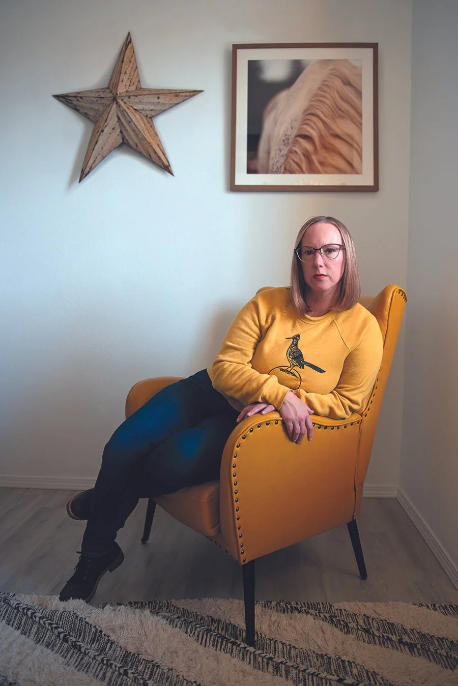 A woman with glasses and shoulder-length hair sitting in a mustard yellow armchair in a room with white walls. She is wearing a yellow sweatshirt with a bird graphic and black pants. There is a wooden star and framed photographs of a close-up of a hedgehog on the wall behind her, and a striped rug on the floor.