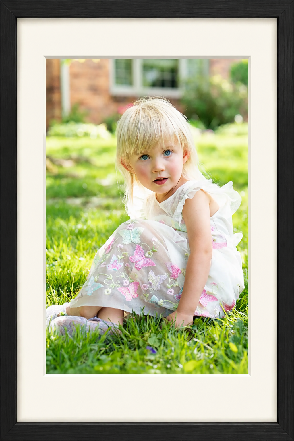 Portrait of a young girl with blonde hair and blue eyes sitting on grass outside, wearing a white dress with pink and blue butterfly embroidery.