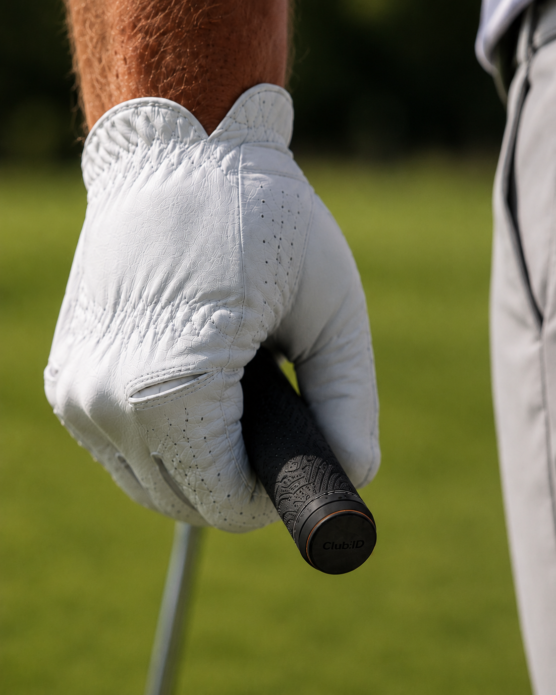 Close-up of a golfer wearing a white golf glove gripping a golf club, with part of another person visible in the background, on a grassy golf course.