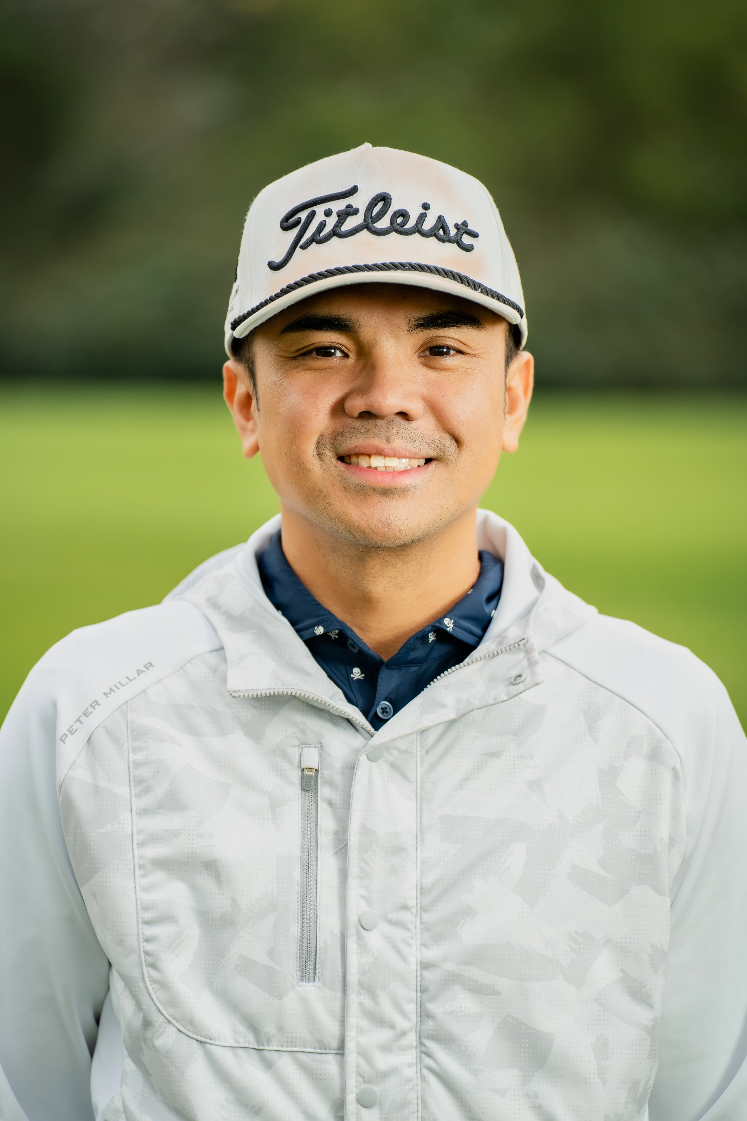 A man smiling outdoors, wearing a white golf cap with the word "Titleist" embroidered on it, a white jacket, and a navy blue shirt with small white skull print, on a golf course with blurred green trees in the background.