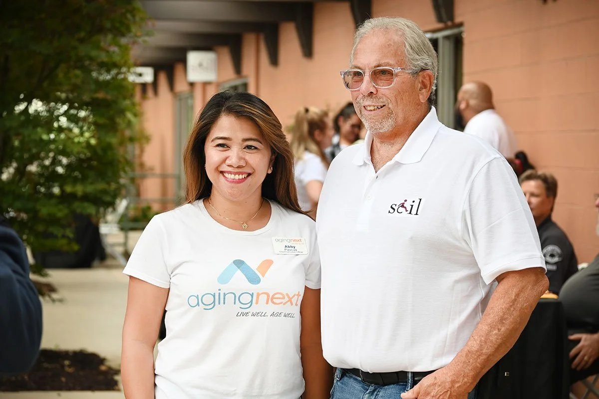 Abby Pascua and Larry Grable stand together outside at a community event, smiling. Abby wears an AgingNext shirt, and Larry wears a Service Center for Independent Life shirt.