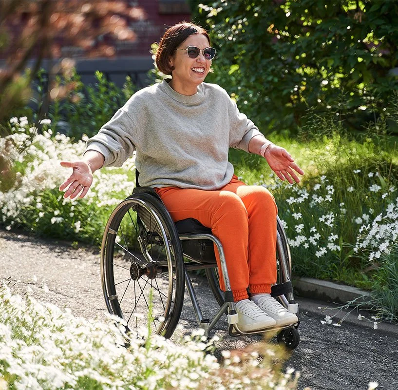 A woman wearing sunglasses and seated in a wheelchair on a garden path smiling with arms outstretched, surrounded by green plants and white flowers on a sunny day.