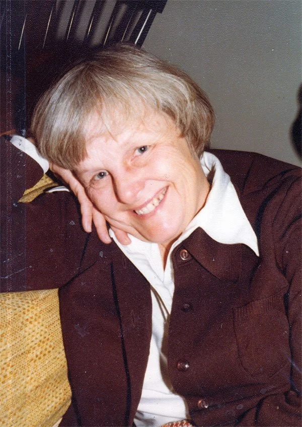 Mary Sandoe smiles at the camera while resting her cheek on her hand. She sits on a yellow patterned couch and wears a dark sweater over a white collared shirt.