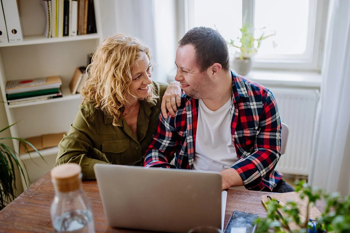A woman rests her hand on a man’s shoulder as they smile at each other over a laptop. They sit at a wooden table in a bright room with shelves and a window.