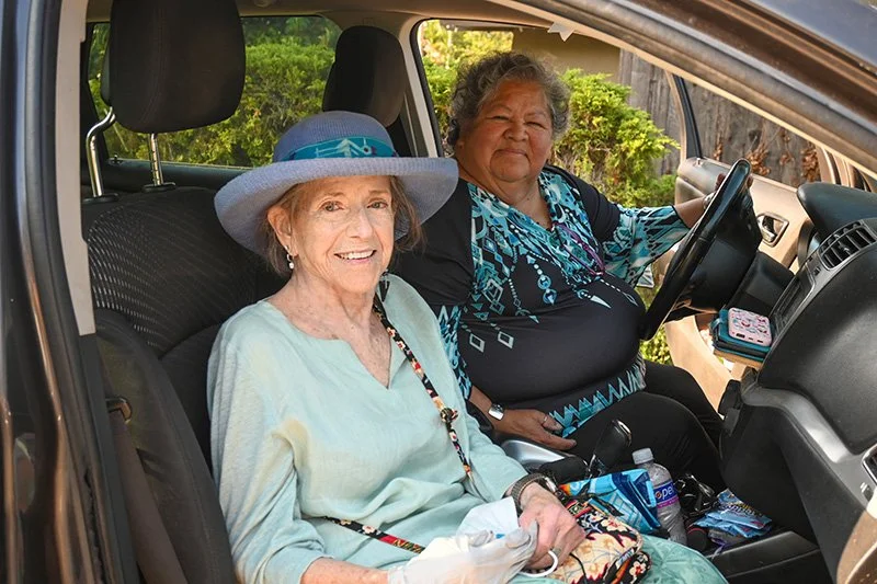 Two older women smile from the front seats of a parked vehicle. The passenger wears a wide-brimmed blue hat. Outside the vehicle it is sunny and bright, with green foliage visible through the windows.