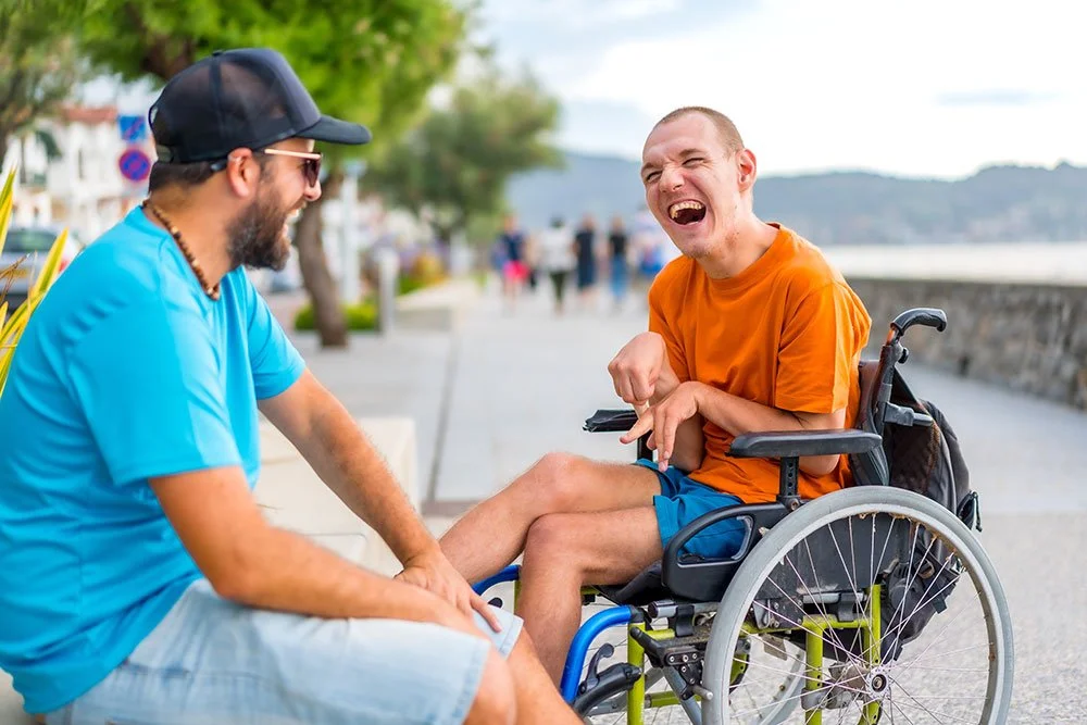 A man in a wheelchair laughs with a companion seated beside him on a sunny beachfront walkway. Trees, water, and distant hills appear behind them.