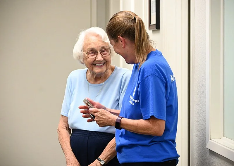 A volunteer in a blue shirt talks with an older woman. The older woman smiles as they stand together near a doorway.