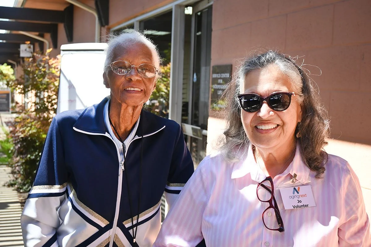 Two women smiling outside a building, one with gray hair and glasses wearing a navy jacket, and the other with curly hair, sunglasses, and a pink shirt with a volunteer badge.