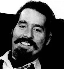 A black-and-white close-up portrait of Ed Roberts smiling. He has dark hair, a mustache, and a beard.