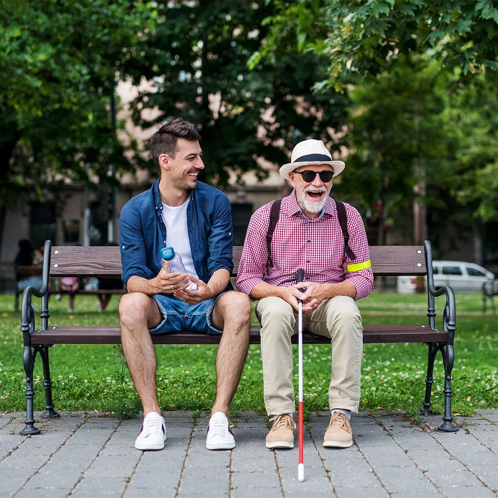 A visually impaired older man and a younger man sit together on a park bench, smiling and laughing. The older man holds a white cane and wears sunglasses and a sun hat, while the younger man holds a water bottle.