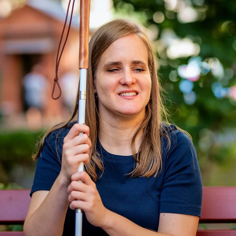 A young, visually impaired woman with light brown hair and wearing a navy blue t-shirt sits on a park bench. She holds a white walking cane and smiles.