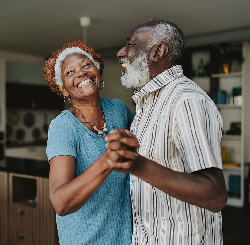 An older couple dance together in their living room, smiling and enjoying each other's company. Family photos are visible behind them.