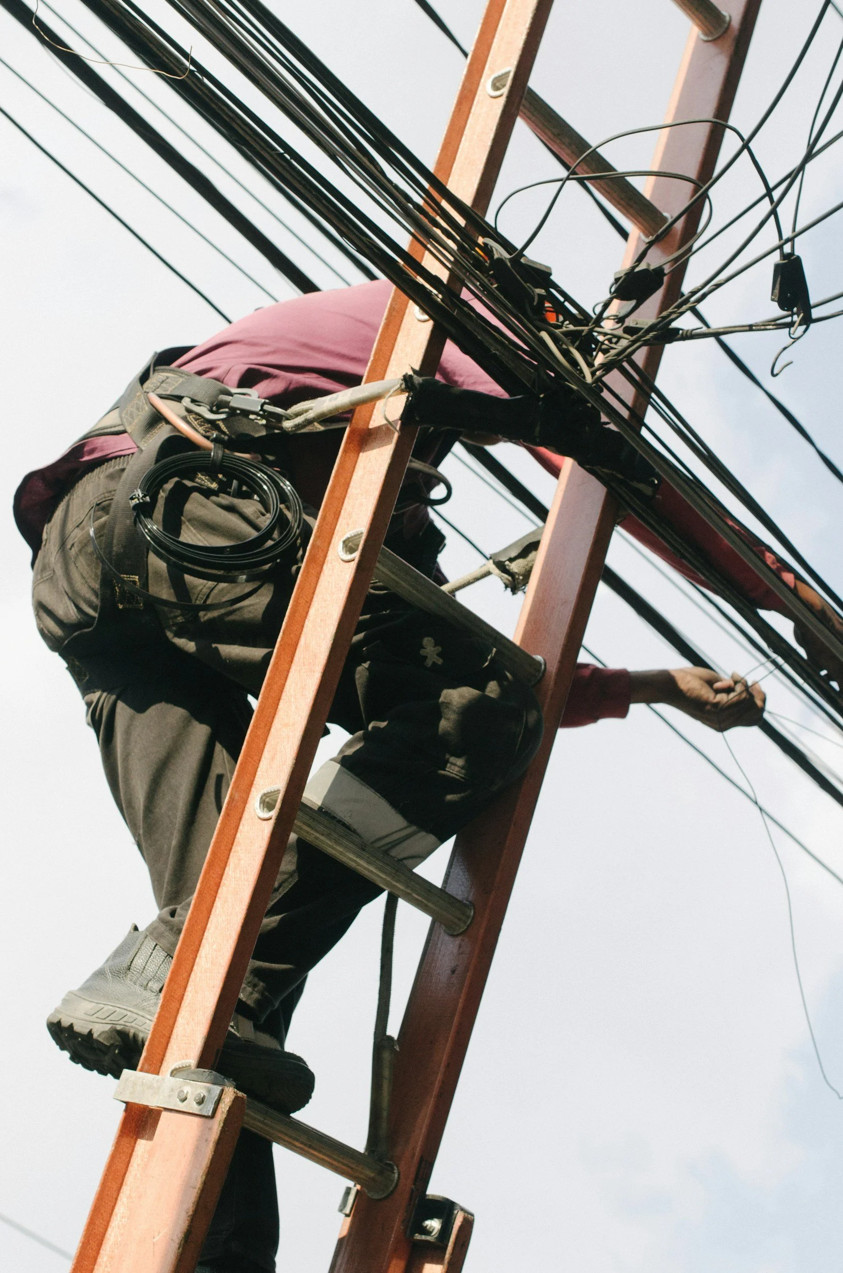 A person working on an electrical pole, climbing a tall wooden ladder with wires attached.