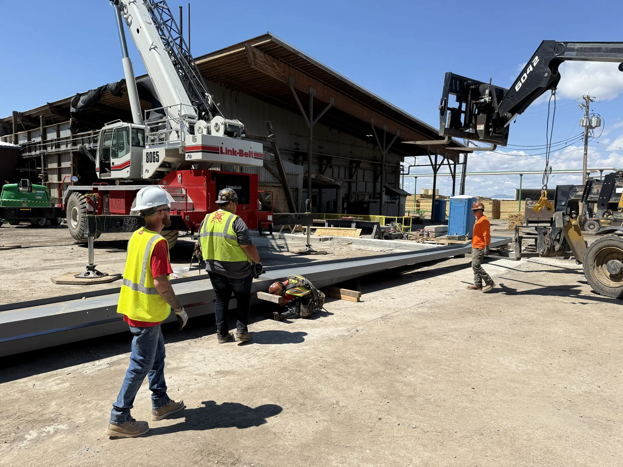 Workers wearing safety vests and hard hats on a construction site, operating large equipment including a crane lifting a steel beam in front of a barn-like structure under a blue sky.