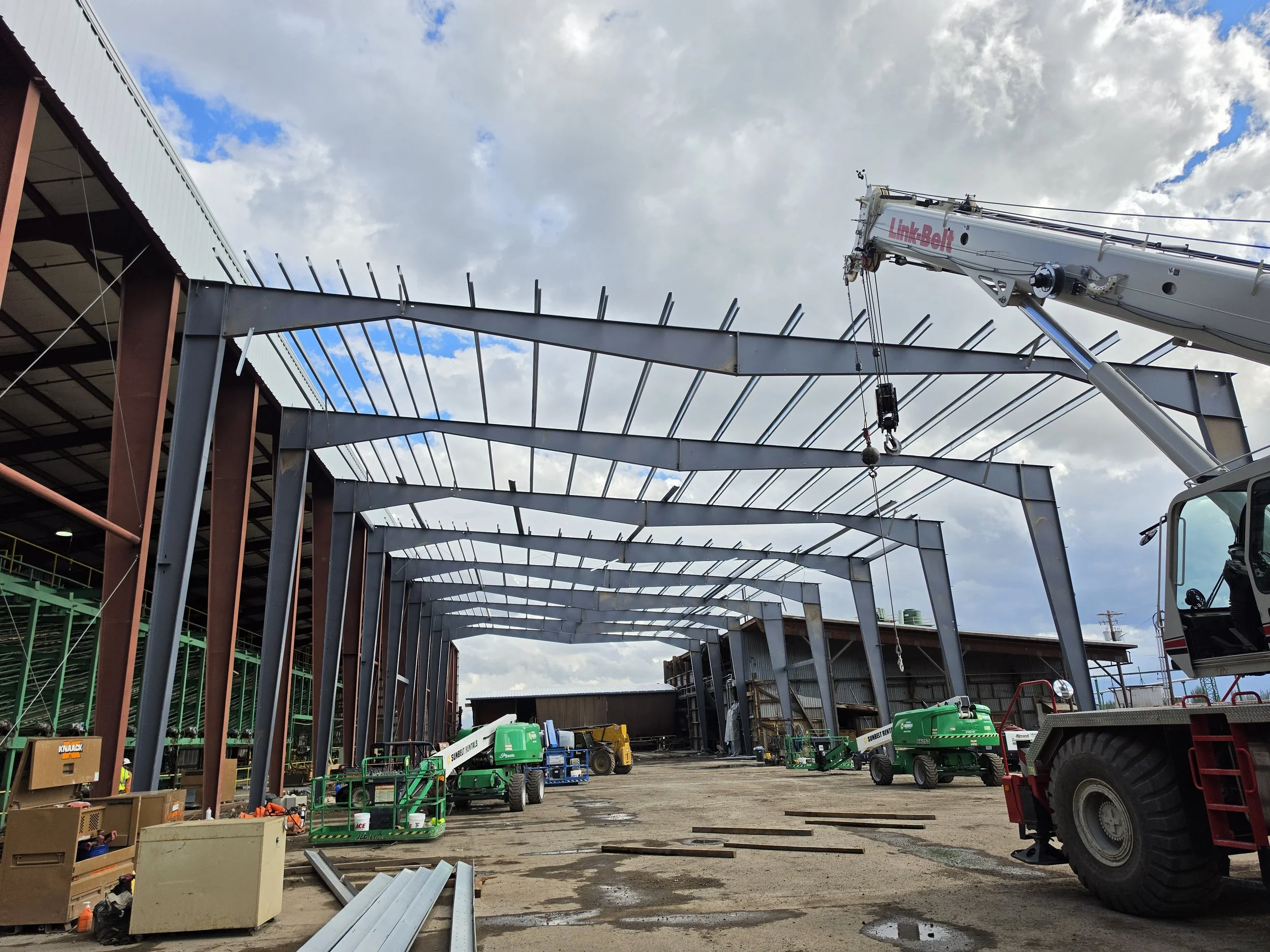 Construction site with steel framework, cranes, construction vehicles, and building materials under a partly cloudy sky.