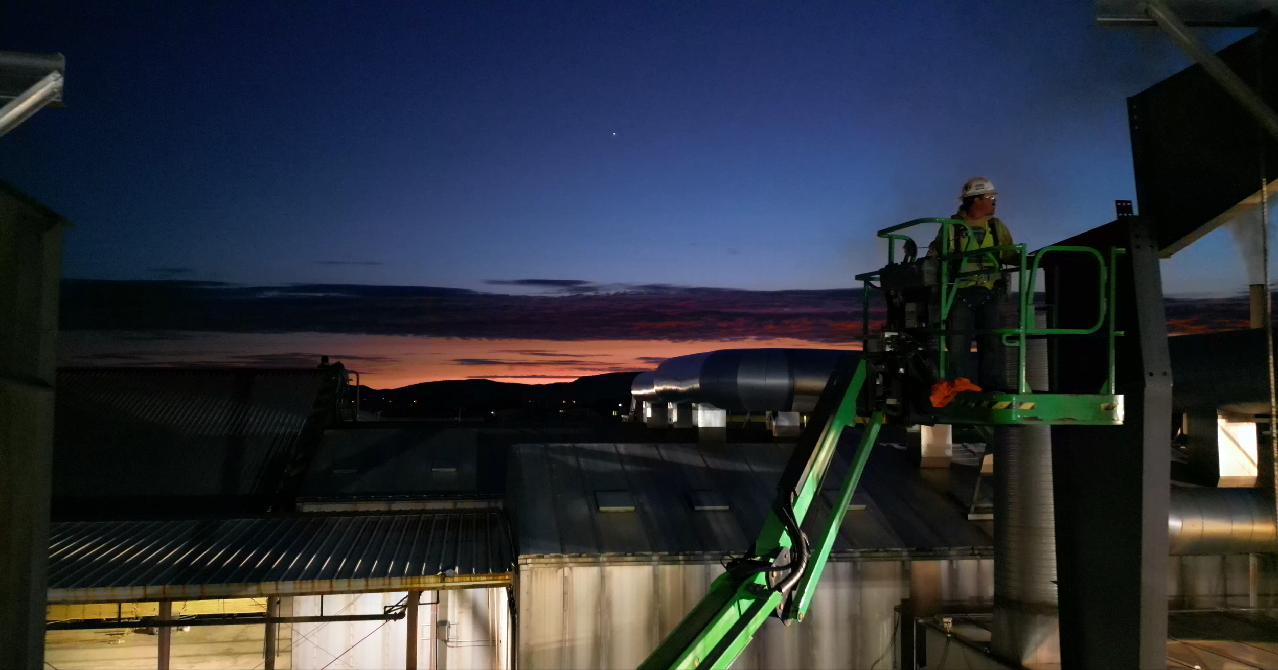 A worker wearing safety gear stands on a green lift platform inspecting equipment on industrial rooftops during sunset or sunrise, with a colorful sky and hills in the background.