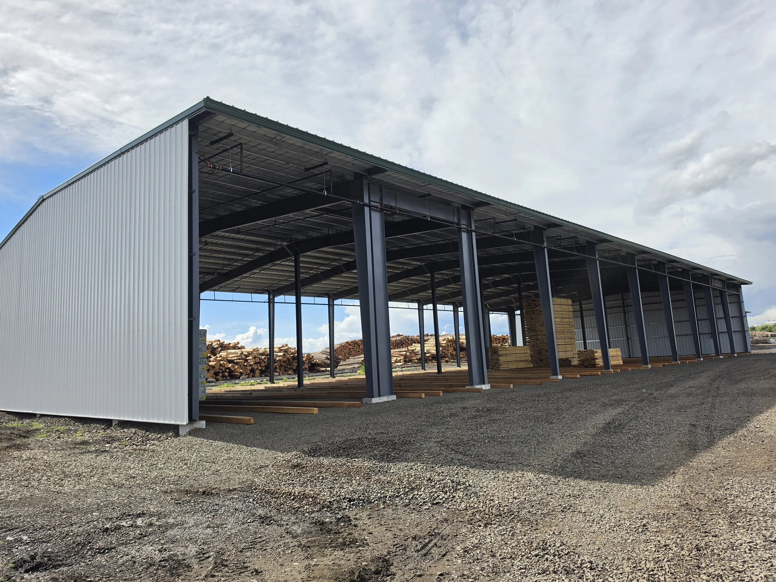 A large open-sided metal building under construction with logs stacked outside underneath cloudy skies.