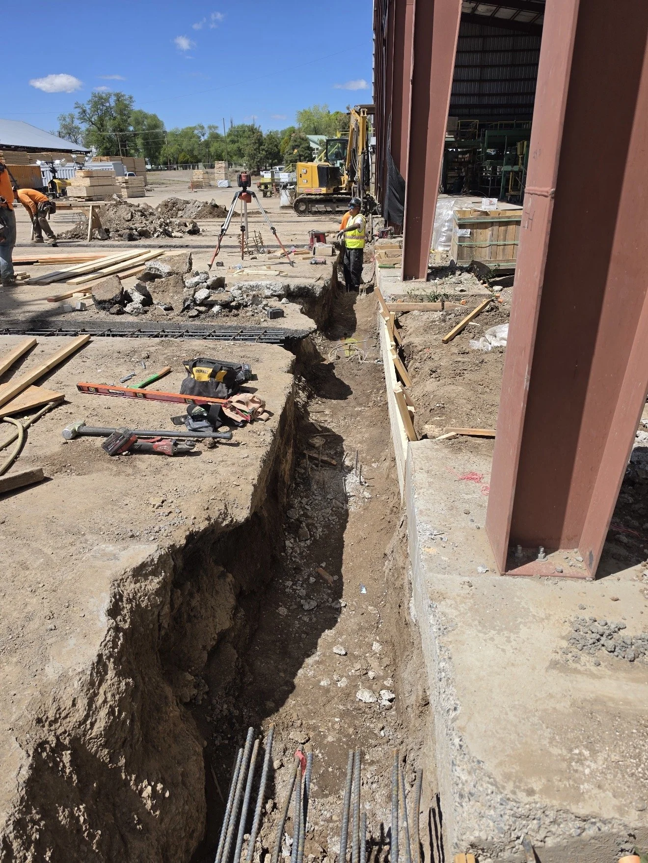 Construction site with workers and machinery, trench excavation for foundation, building materials scattered around, and a clear blue sky.