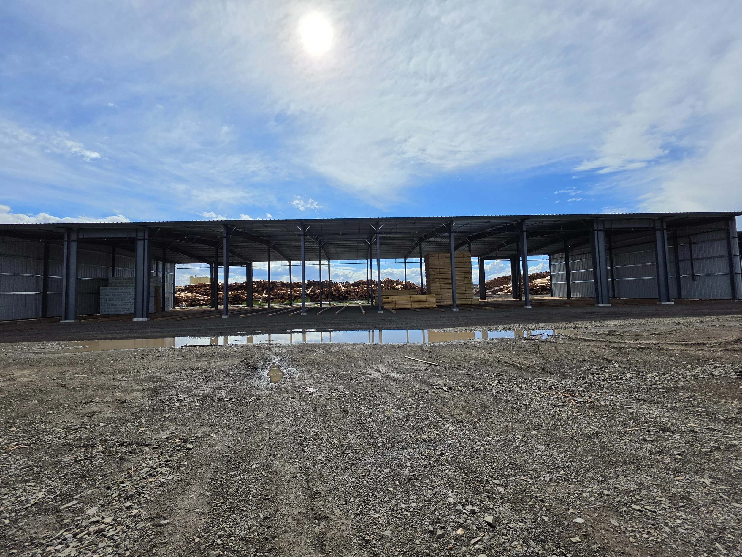 A partially constructed metal building with an open front and a stack of lumber inside. The ground is rough and muddy with puddles, and the sky is mostly cloudy with some blue showing.