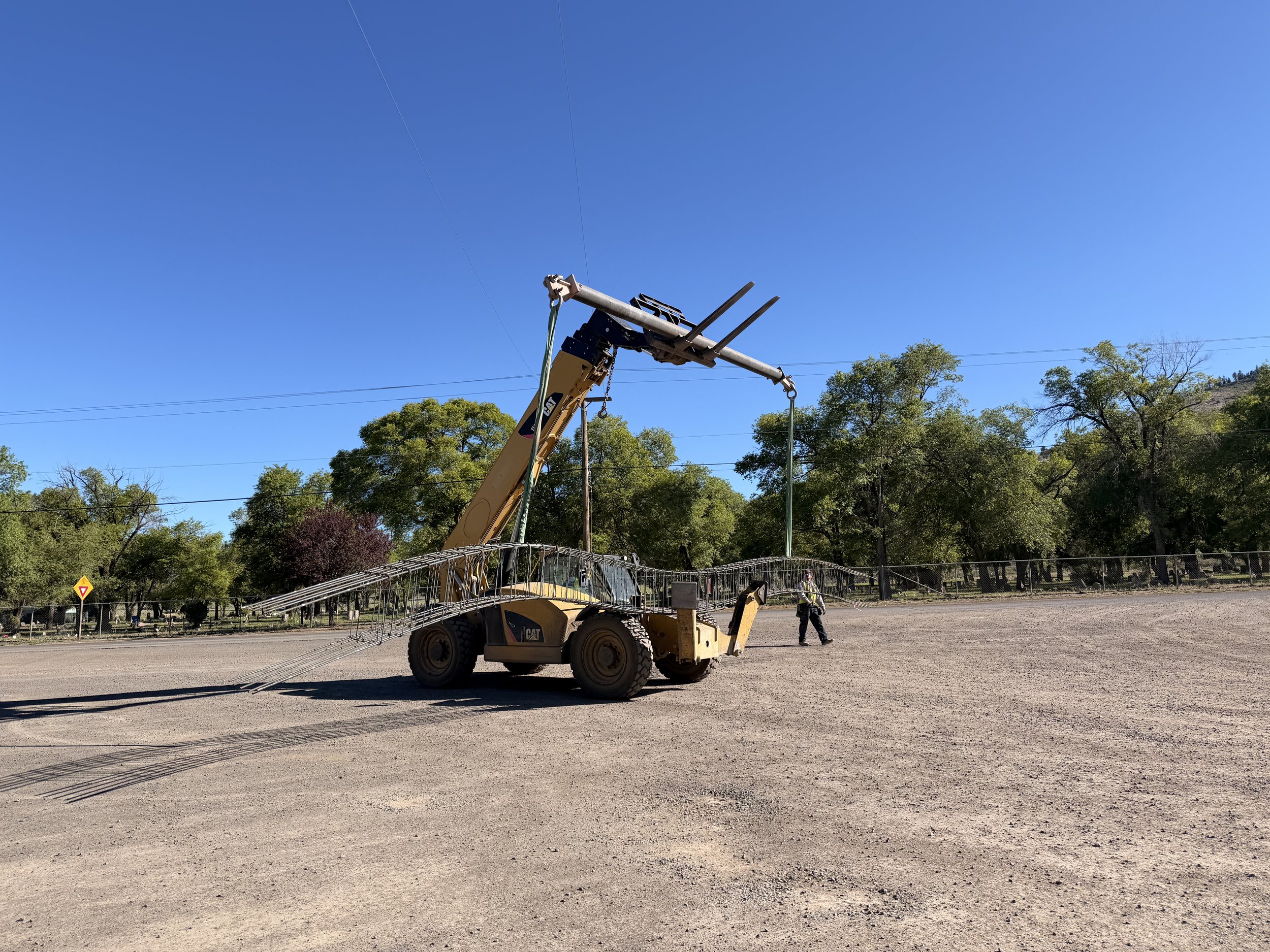 A construction vehicle, possibly a backhoe or excavator, lifting a tall pole in an open area with trees and a blue sky in the background.