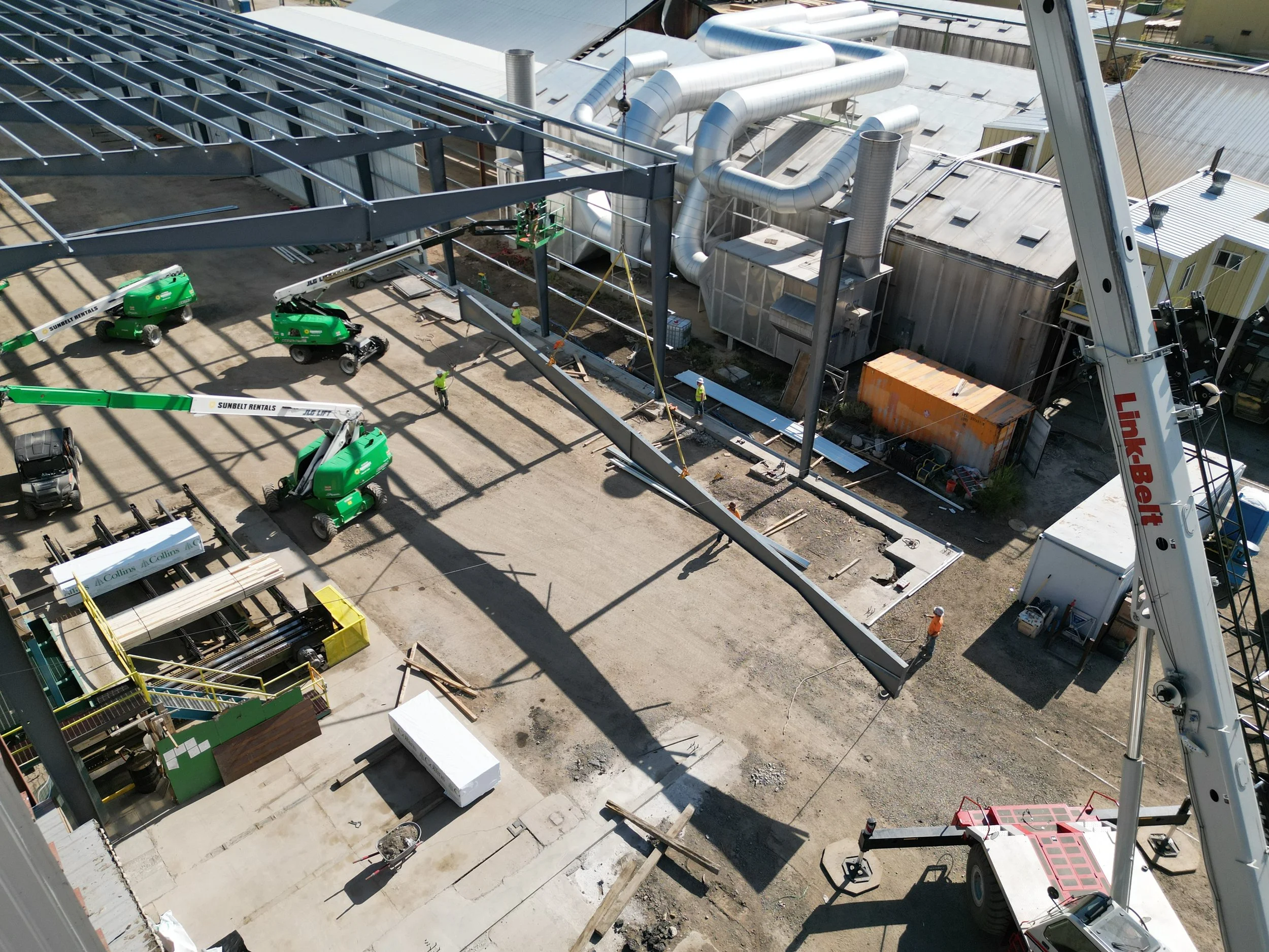 Construction site with steel framework, ductwork, and workers installing building components. Multiple green lifts and a crane are visible.