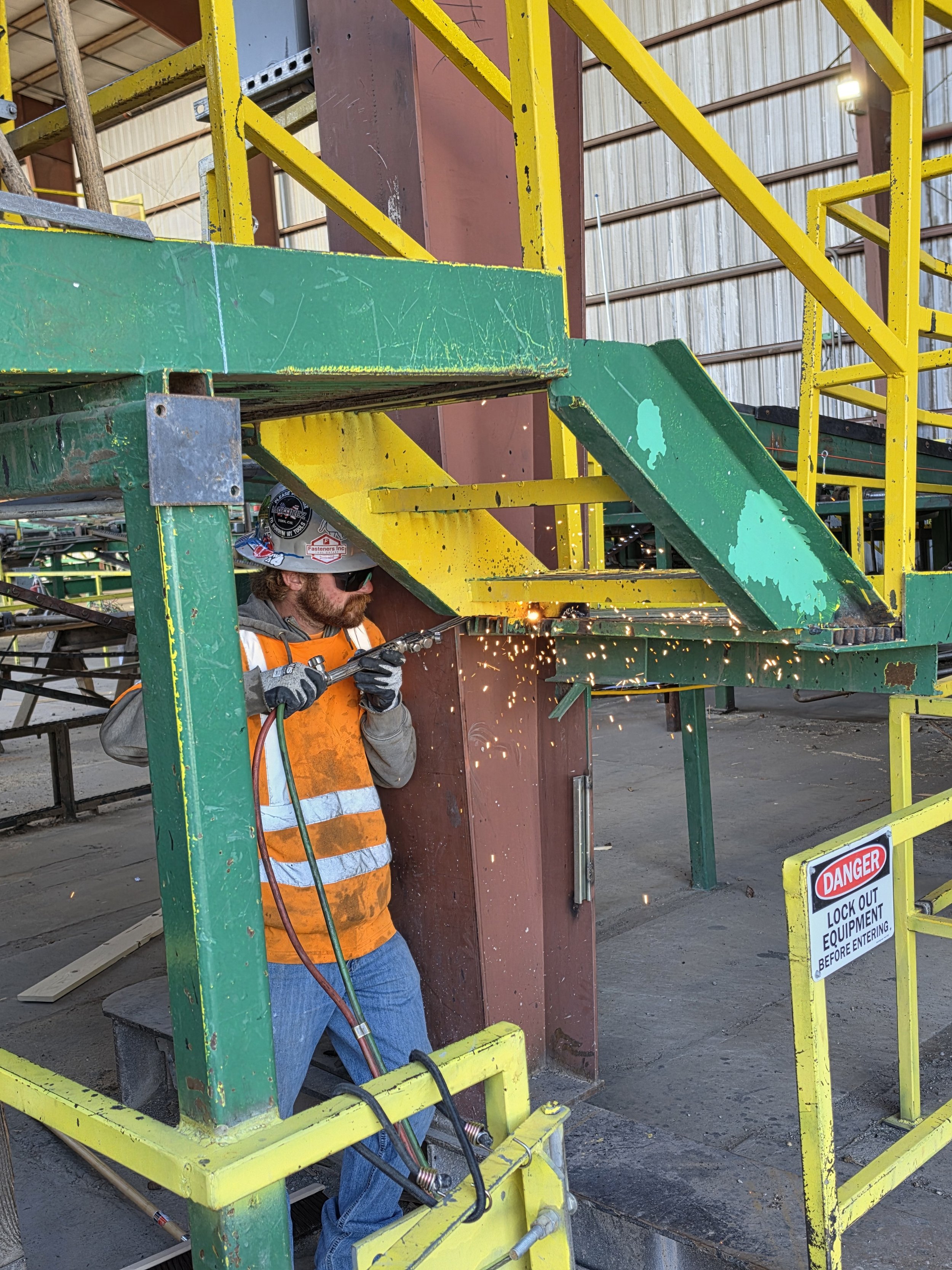Worker welding metal beams inside a yellow safety enclosure at a construction site, with sparks flying and a warning sign that reads 'Danger Lock Out Equipment Before Entering'.