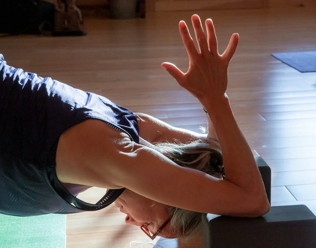 A woman with a headband practicing yoga on a yoga mat in a room with wooden flooring, her right arm raised and her hand open.