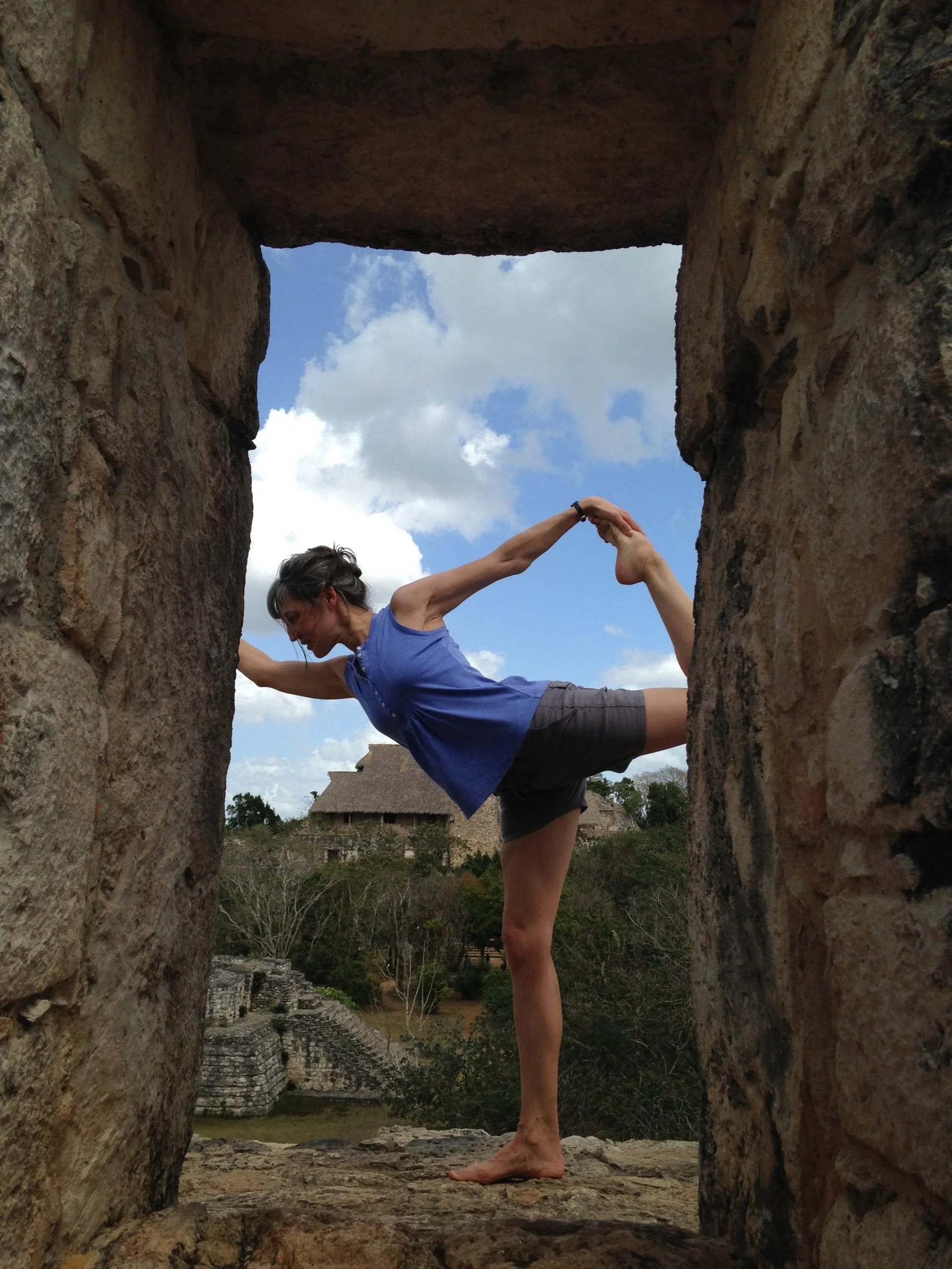 A person standing on one leg in a yoga pose, holding their foot with one hand and the other arm extended, seen through a rectangular opening in a stone wall, with a background of trees and historical buildings under a partly cloudy sky.