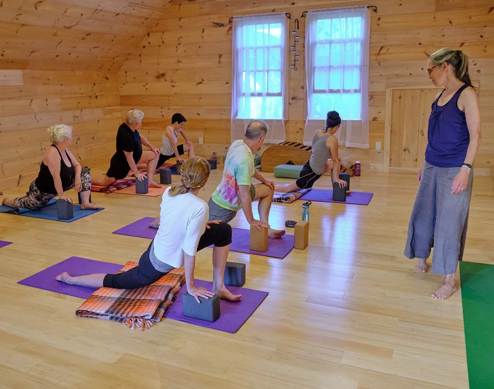 A yoga class with seven participants practicing on yoga mats in a wooden-finished room, guided by an instructor standing nearby.