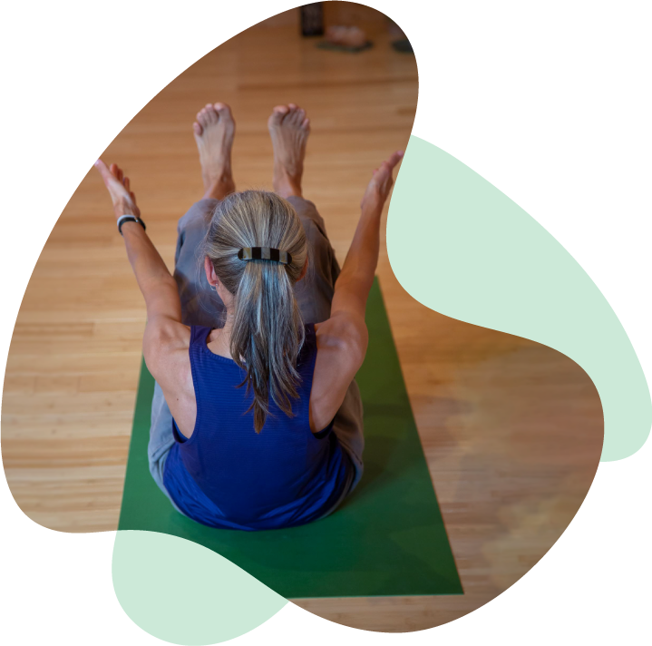 A woman with gray hair tied back, wearing a blue tank top and gray pants, practicing yoga on a green mat in a room with wooden floors.