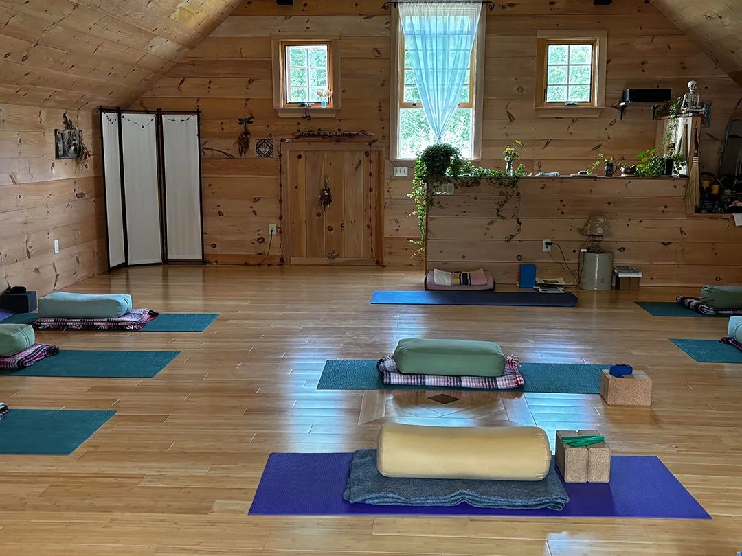 An indoor yoga studio with wooden walls and floor, six yoga mats with bolsters and blocks, and a small windowed wall with plants and decorative items.