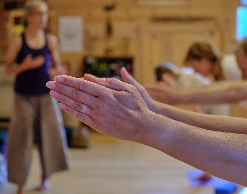 A person practicing yoga in a prayer position, in front of a teacher leading the session in a wooden-finished room.
