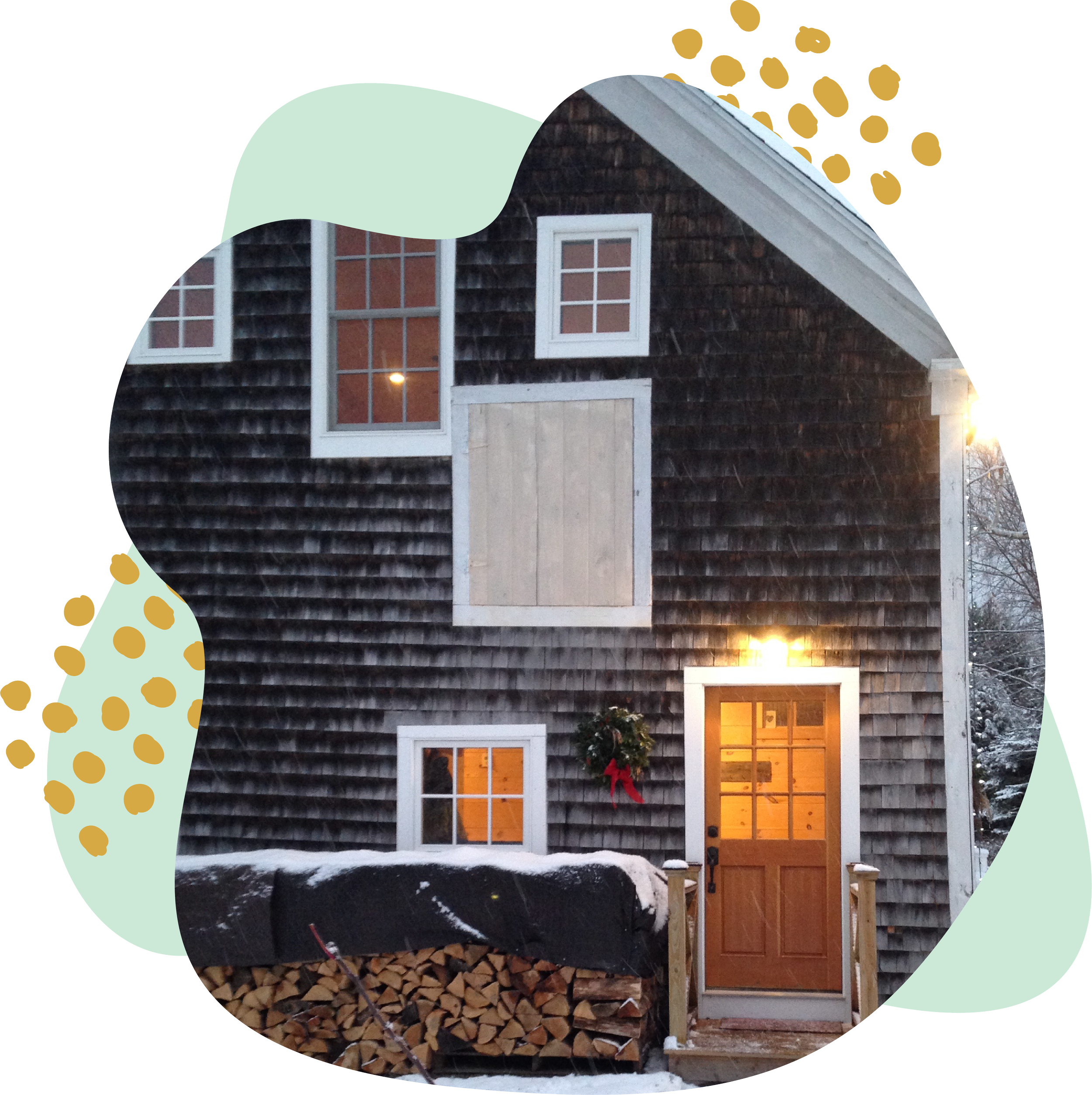 A house with wood siding, white-framed windows, a front door with a Christmas wreath, a pile of firewood, and an outdoor light.