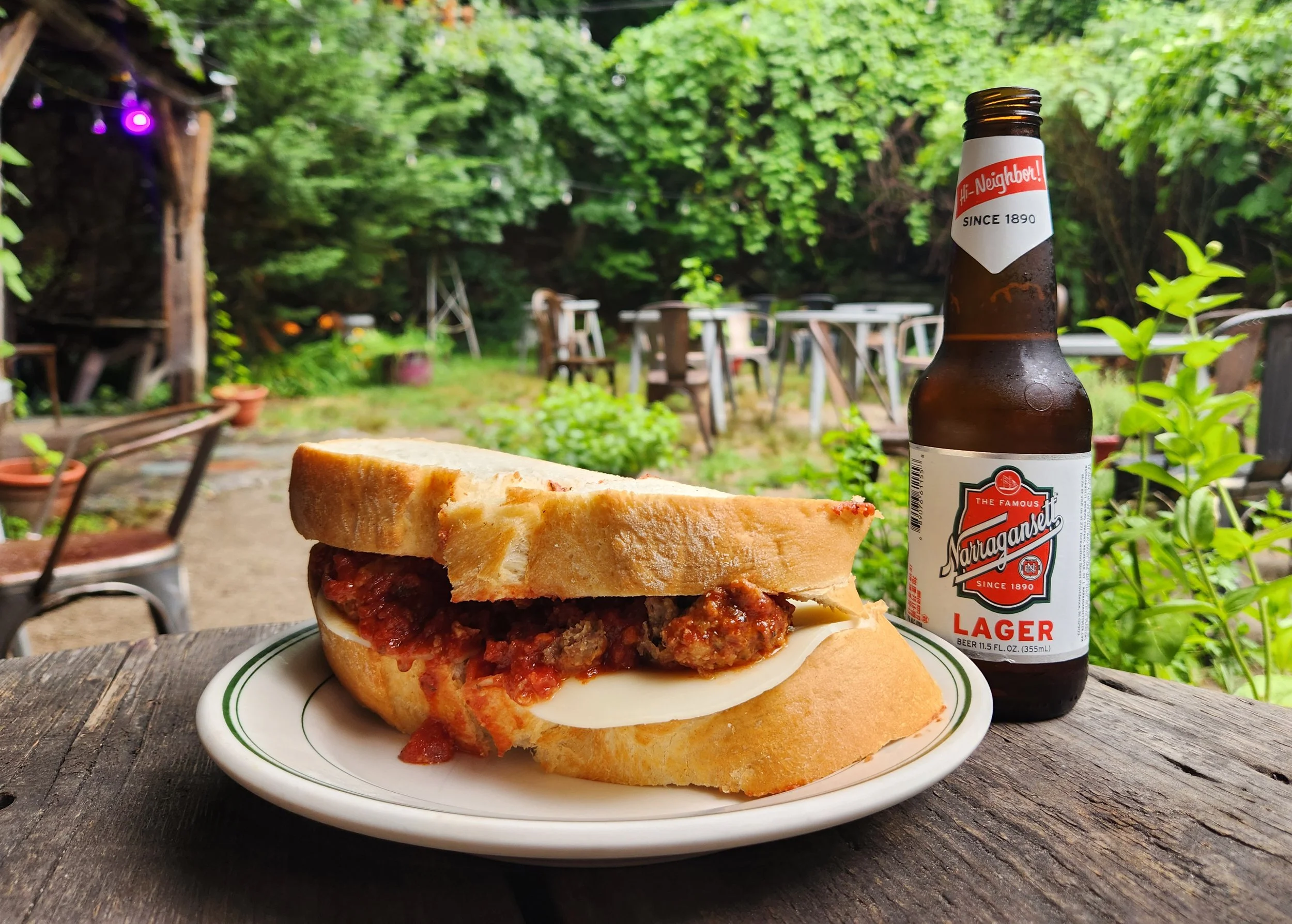 A plate with a sandwich containing ground meatballs, cheese, and tomato sauce, next to a bottle of Narragansett lager beer. The setting is outdoors on a rustic wooden table with a lush green backyard in the background.