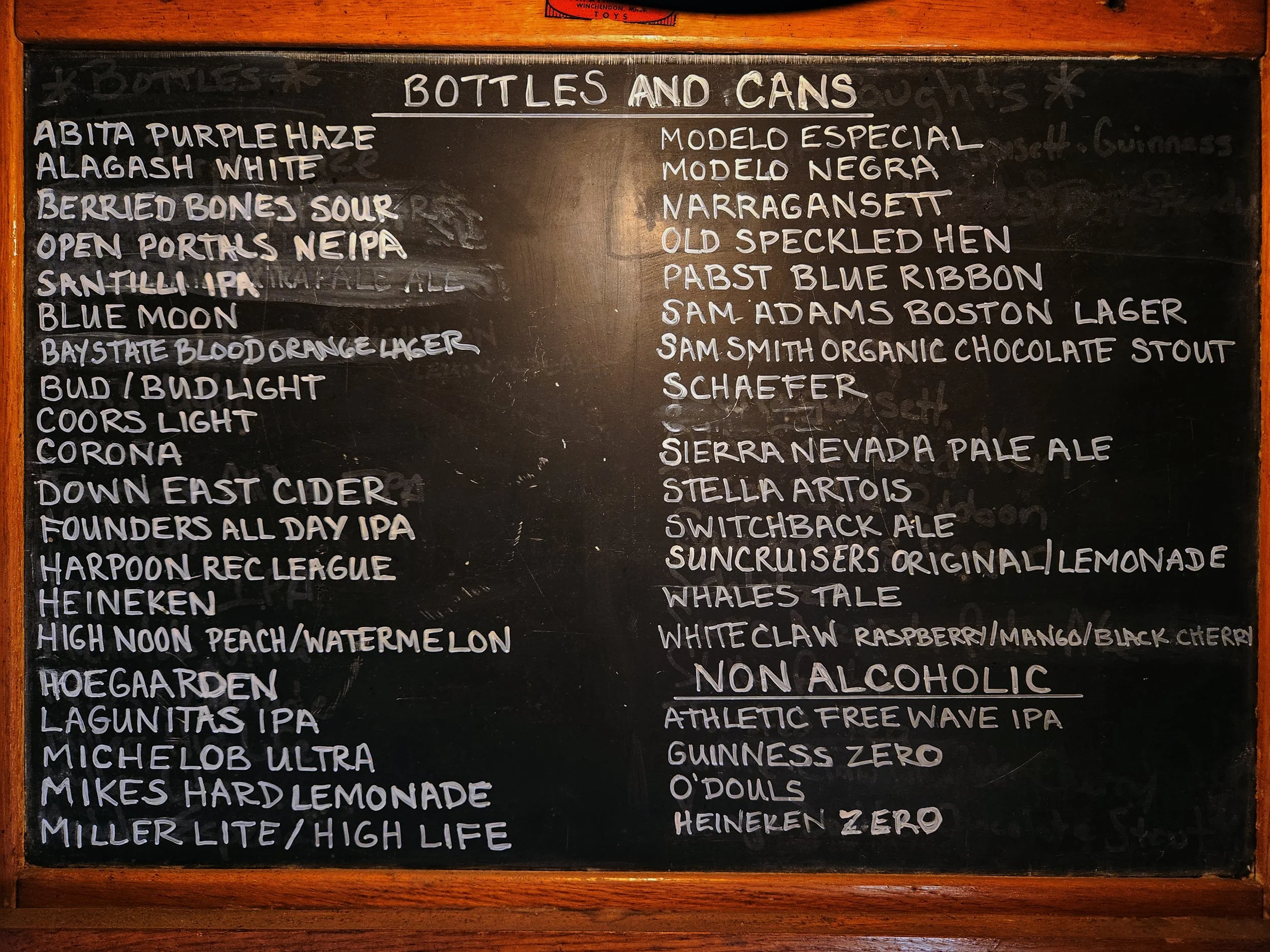 A blackboard menu listing various beers on tap, divided into two columns with the title 'Bottles and Cans' at the top. A large variety of beers and hard drinks are offered, along with a small selection of non-alcoholic beers.