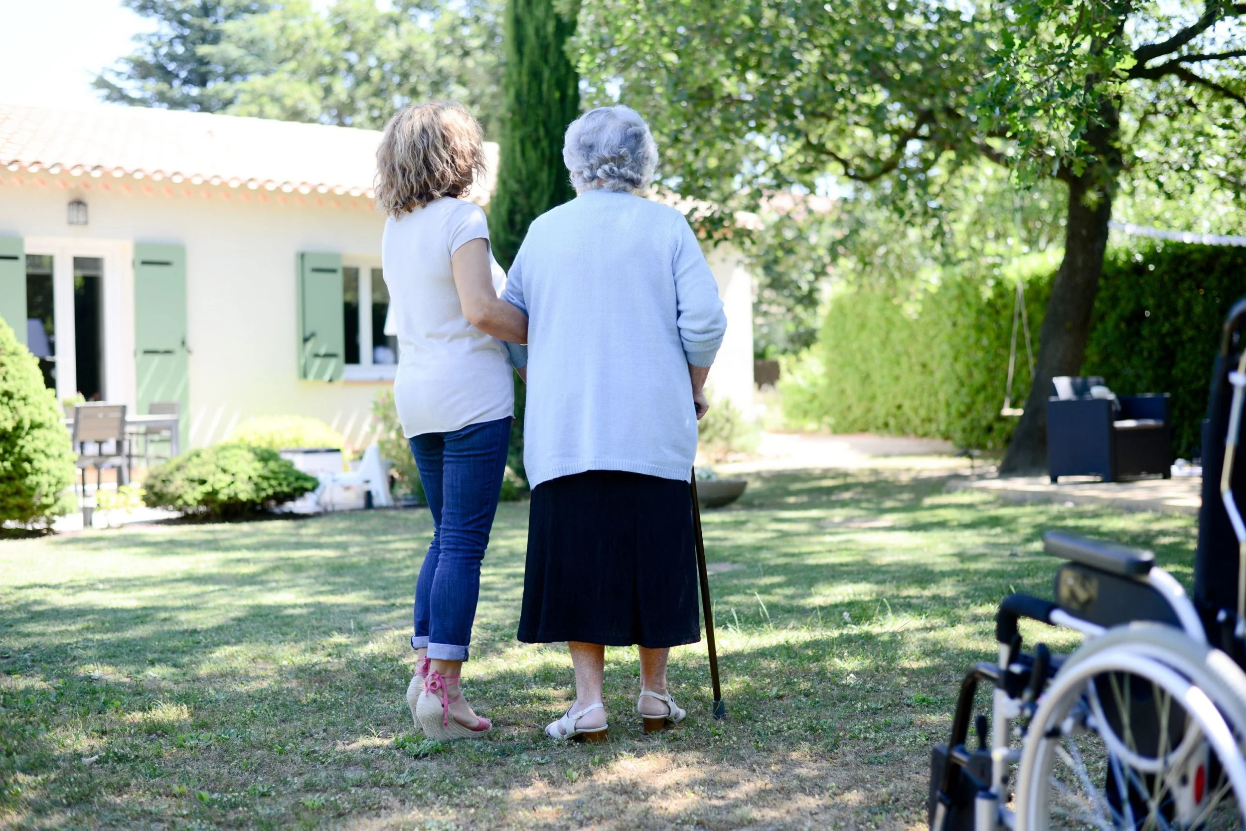 Deux femmes, une jeune et une âgée, se tenant par le bras dans un jardin ombragé, l'une aide l'autre, signe de soutien et de complicité.
