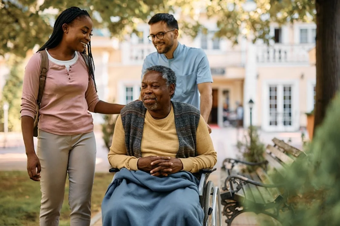 Trois personnes, un homme en fauteuil roulant, une femme et un homme, souriants, dans un parc avec des arbres, en train de discuter et de se soutenir mutuellement.