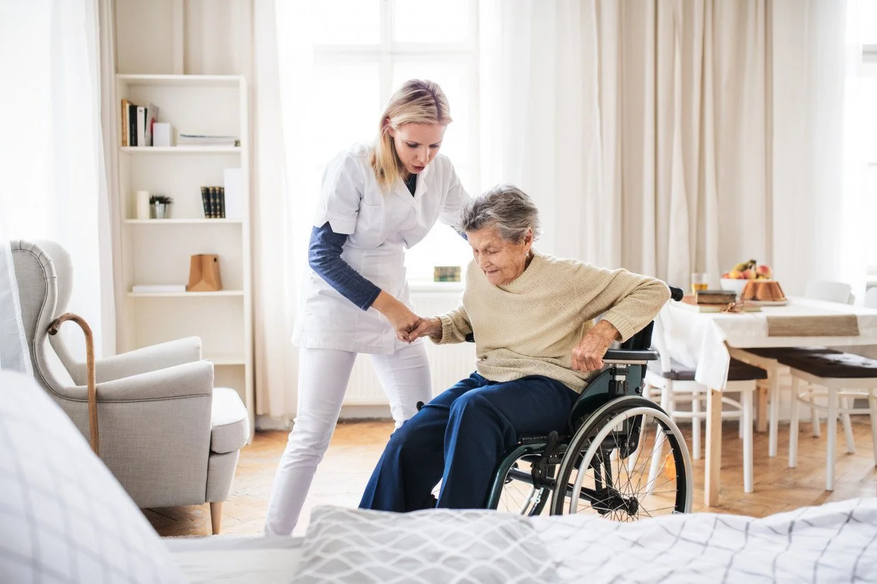 Une infirmière aide une femme âgée en fauteuil roulant dans une chambre lumineuse.