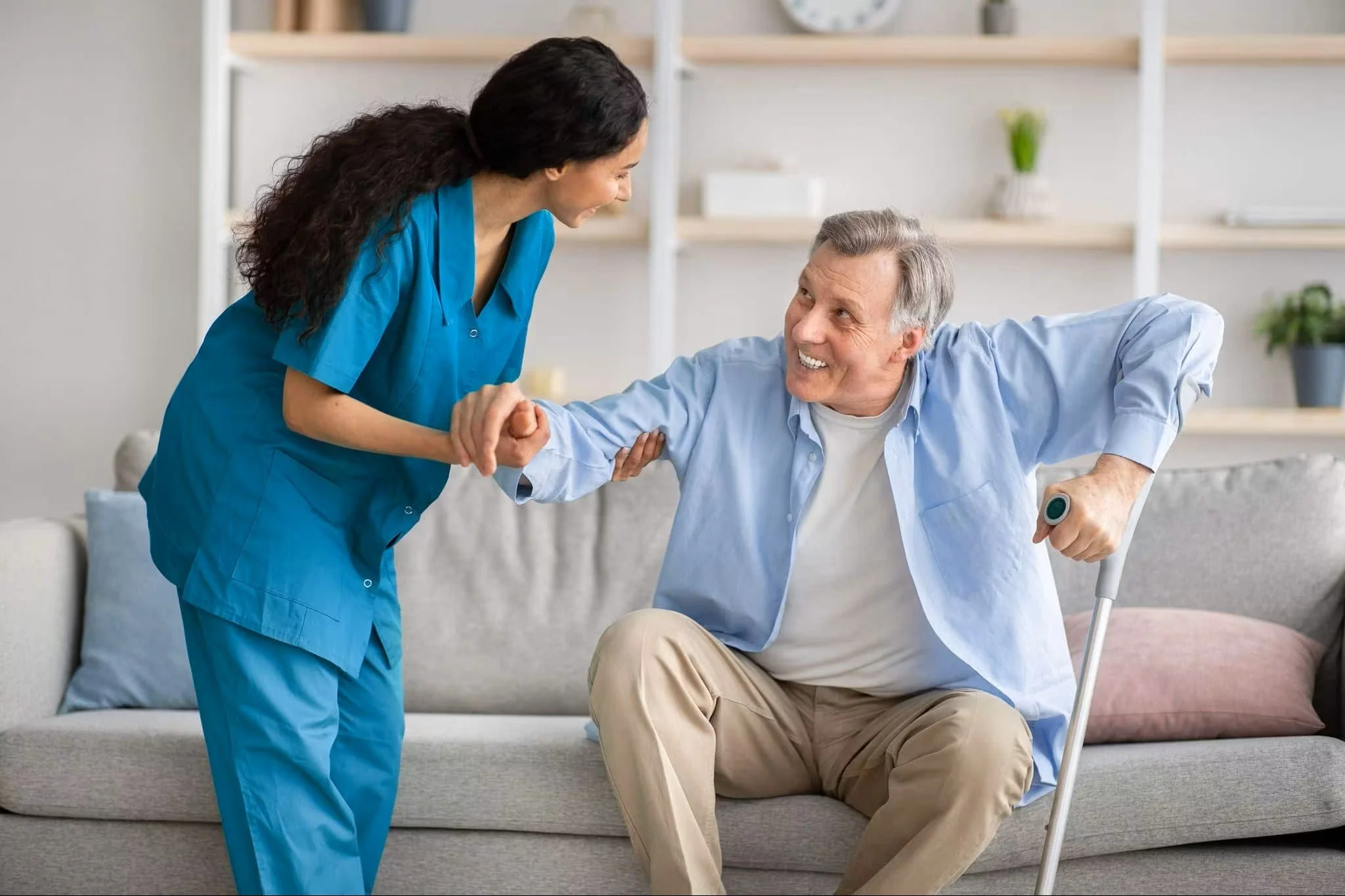 Une infirmière souriante aide un homme âgé à se relever en lui tenant la main dans un salon. L'homme, en fauteuil roulant, a une canne à la main. La scène montre un moment de soutien et de compassion.