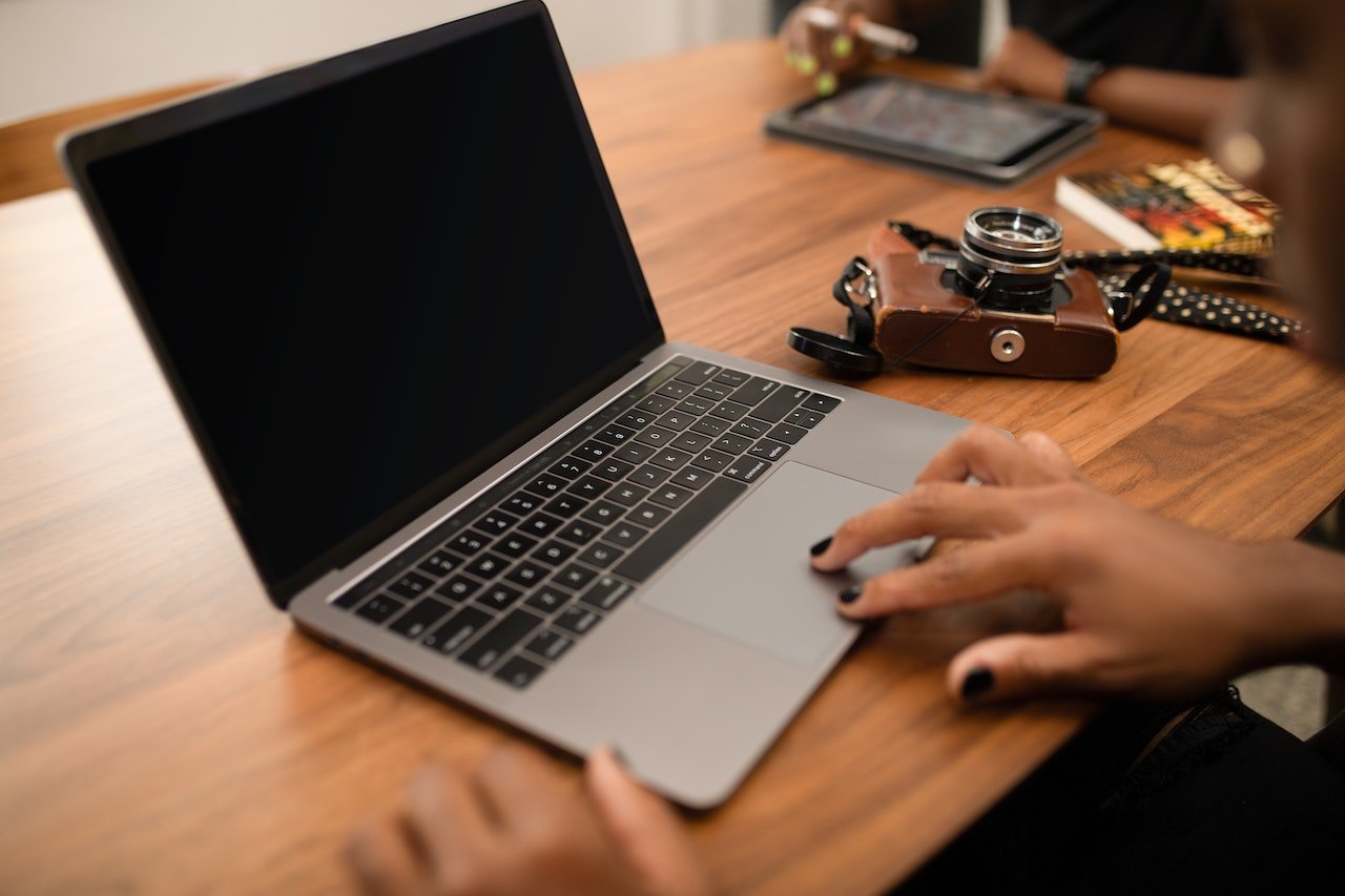 A person using a silver laptop on a wooden table, with a vintage camera and a tablet in the background.