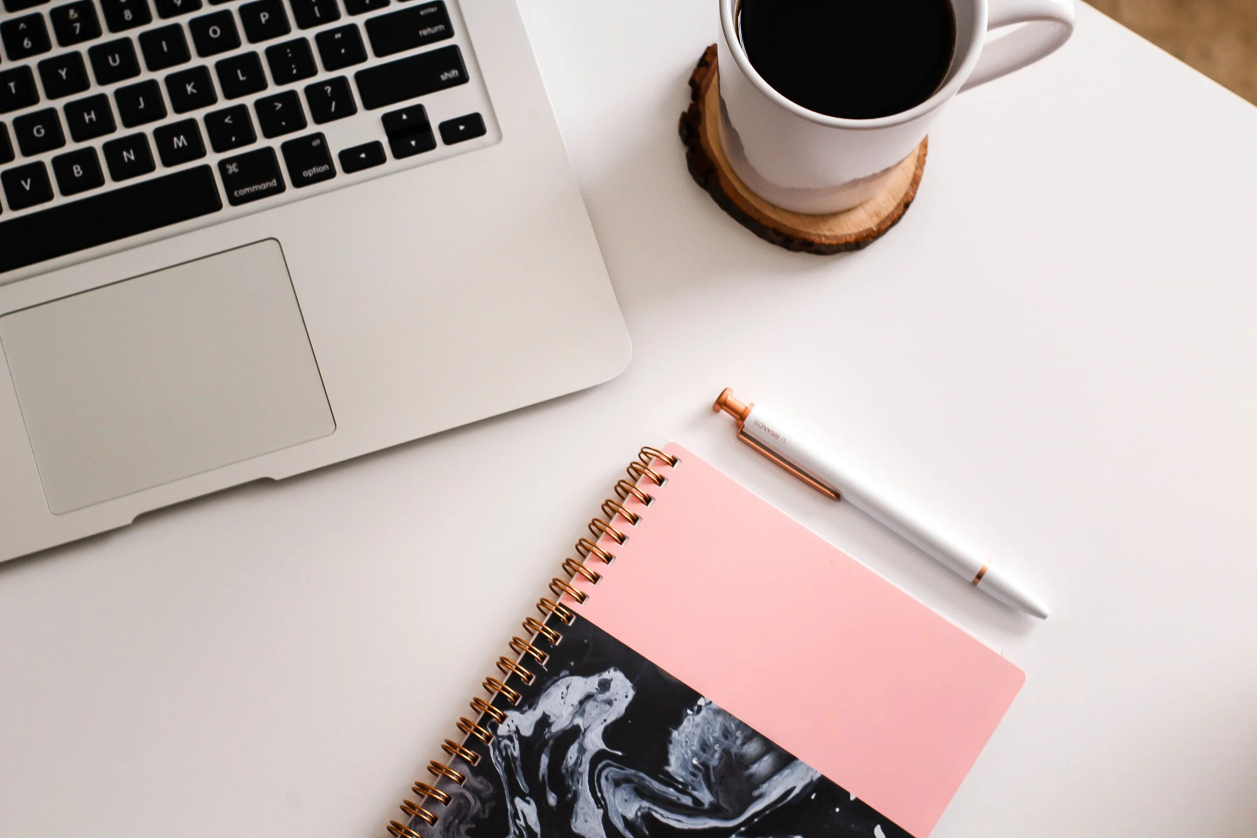 A flat lay of a workspace with a silver laptop, a white cup of coffee on a wooden coaster, a pink spiral notebook, a white pen, and a black and white abstract book on a white surface.