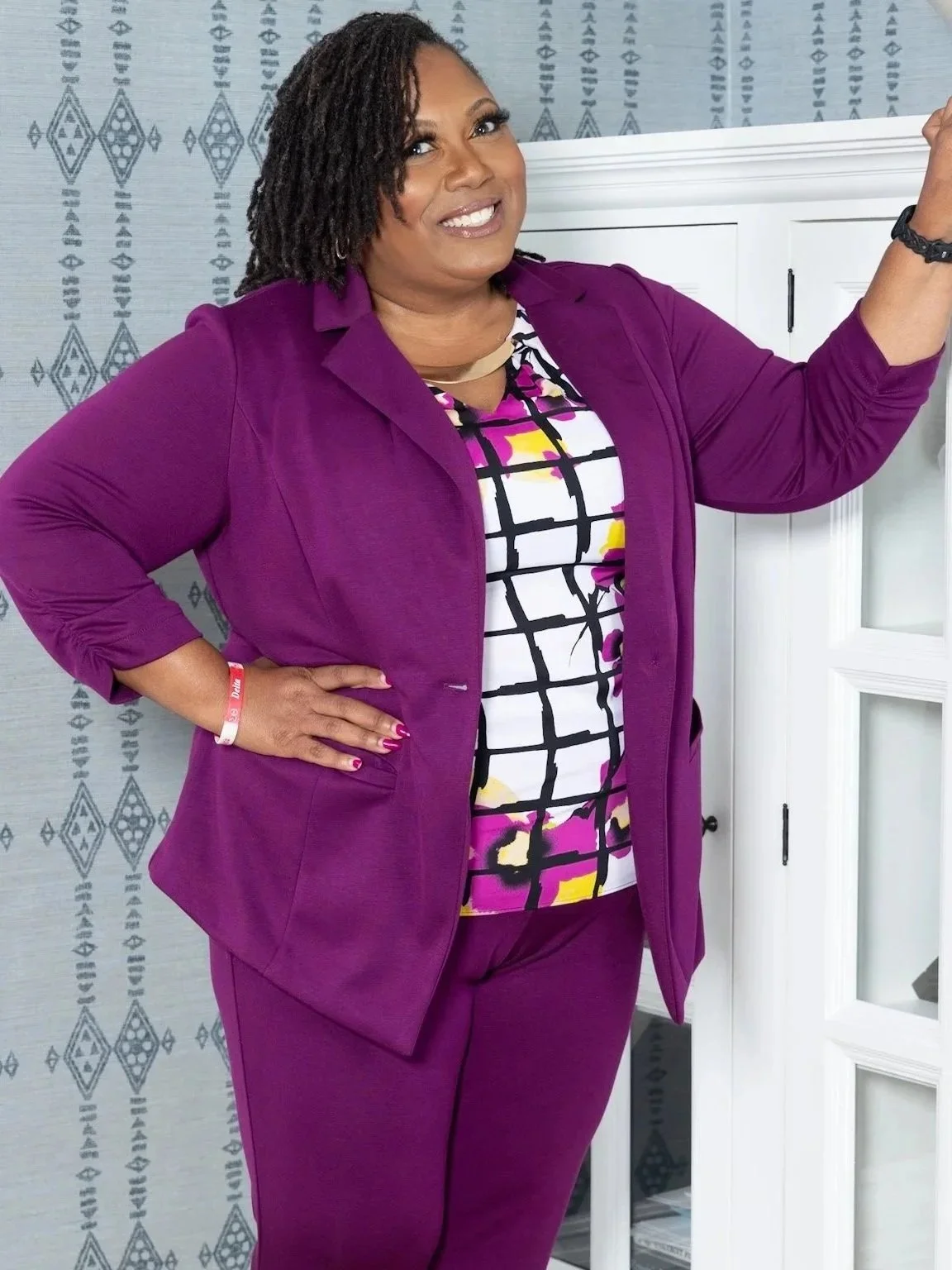A woman with dark curly hair, wearing a purple suit and patterned blouse, standing with one hand on her hip and the other raised, smiling in front of a blue patterned wall and white cabinet.
