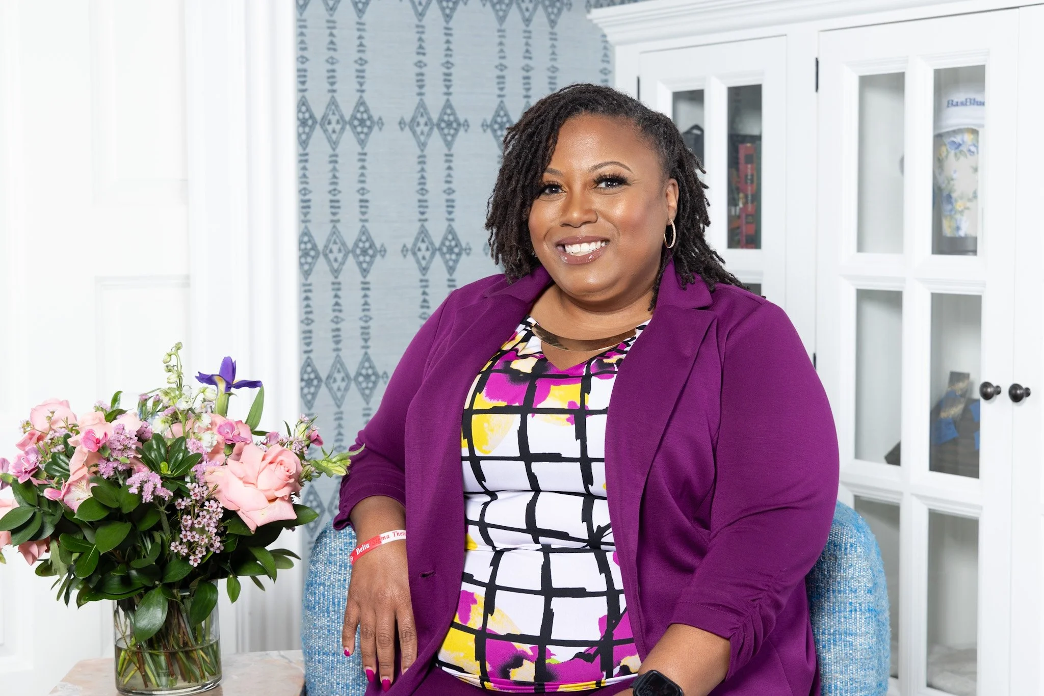 A woman with dark curly hair smiling at a flowers bouquet in a glass vase. She is wearing a purple blazer over a colorful, geometric-patterned dress.
