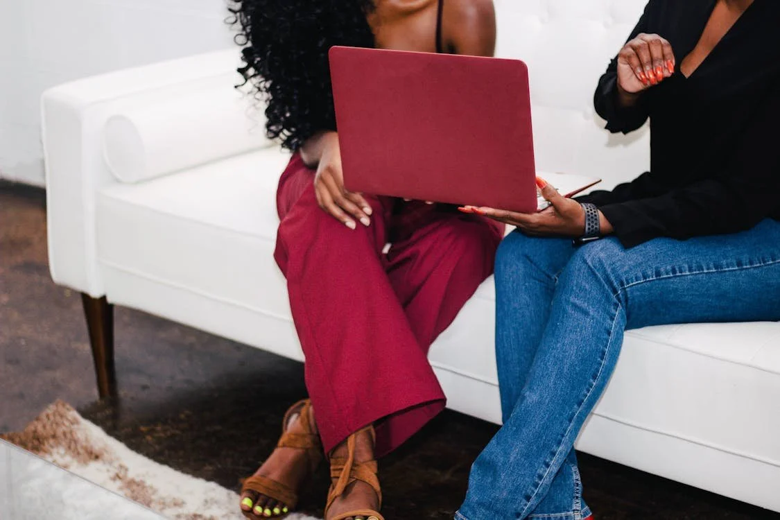 Two women sitting on a white sofa, one showing the other something on a red laptop