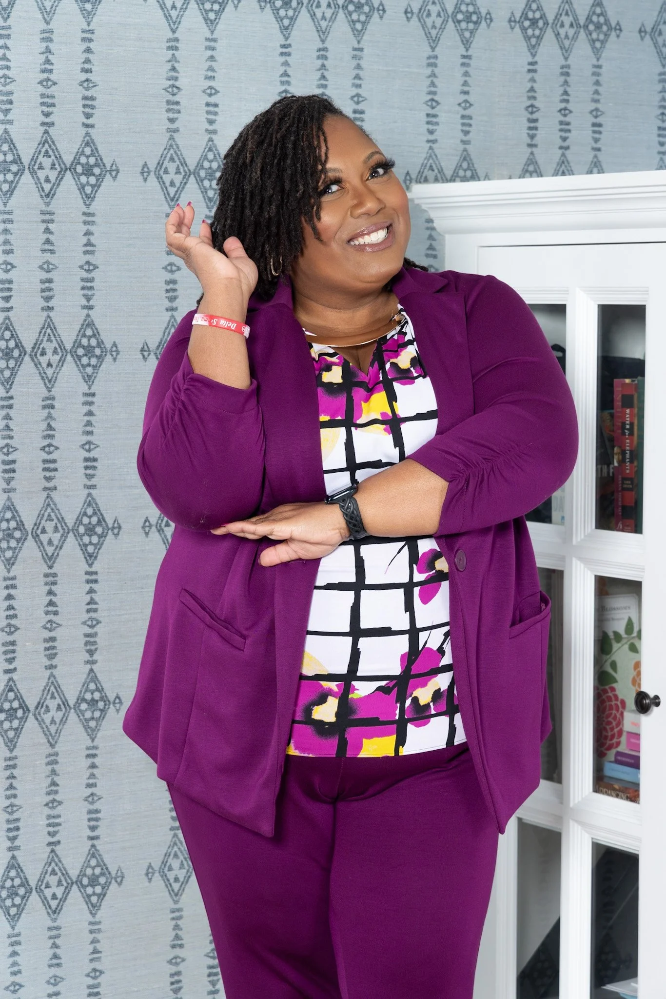A woman with short, curly hair smiling, wearing a purple blazer and purple pants, standing in front of a patterned wall and white cabinet filled with books.